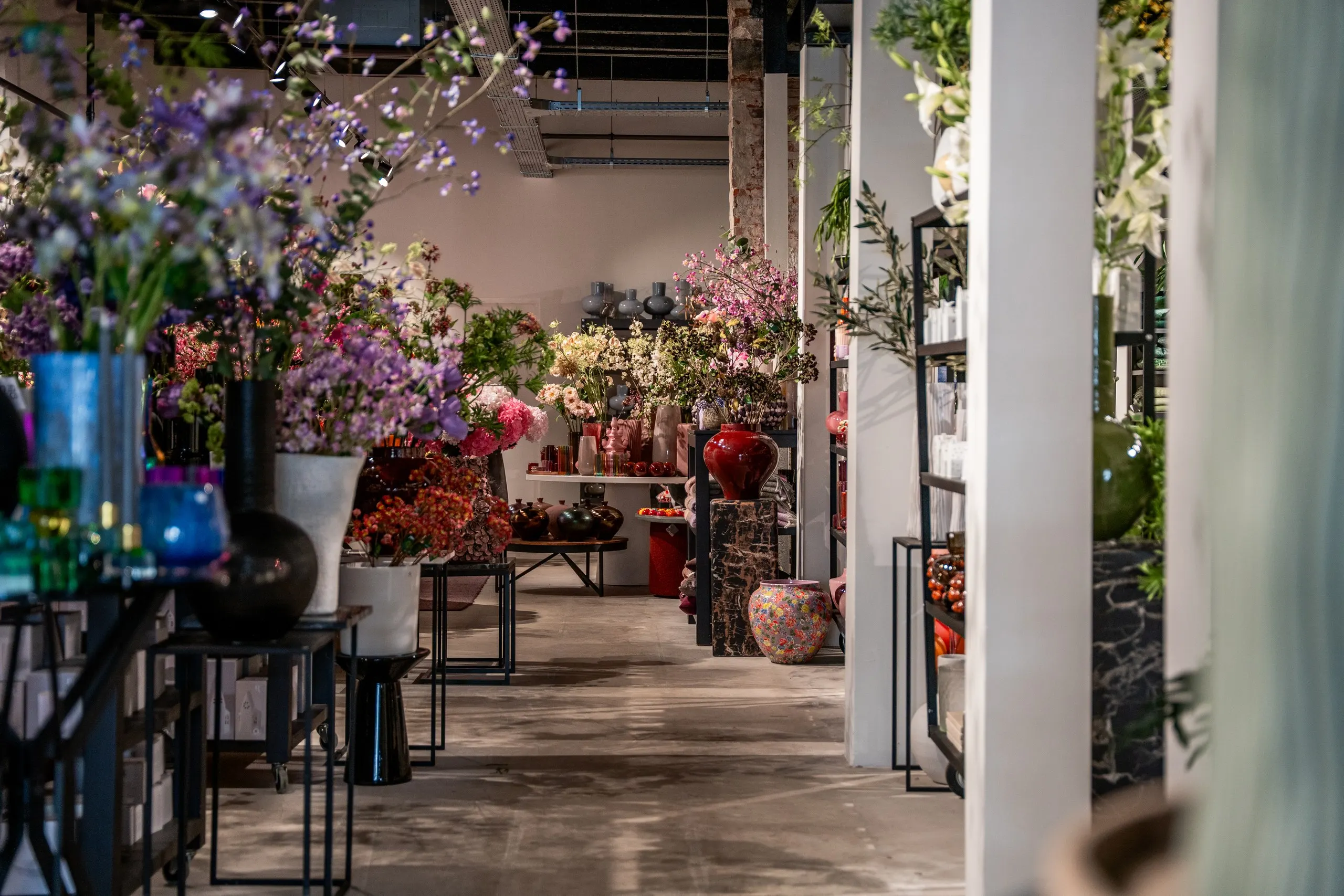 Doorkijk door de winkel: tafels vol kunstbloemen en vazen, een rode pot op een marmeren sokkel en witte pilaren langs het pad