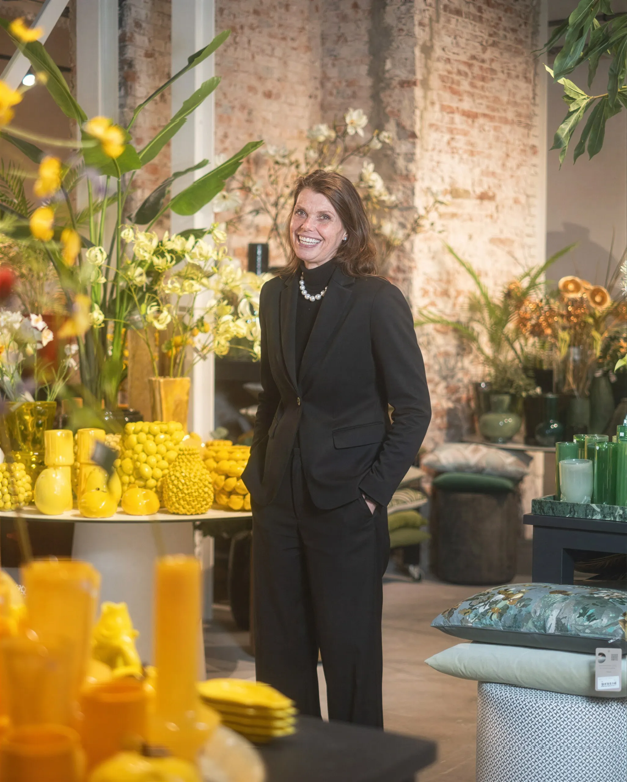 De eigenaresse staat lachend in een zwart pak in de winkel, tussen gele vazen, kunstbloemen en kussens, voor een bakstenen muur