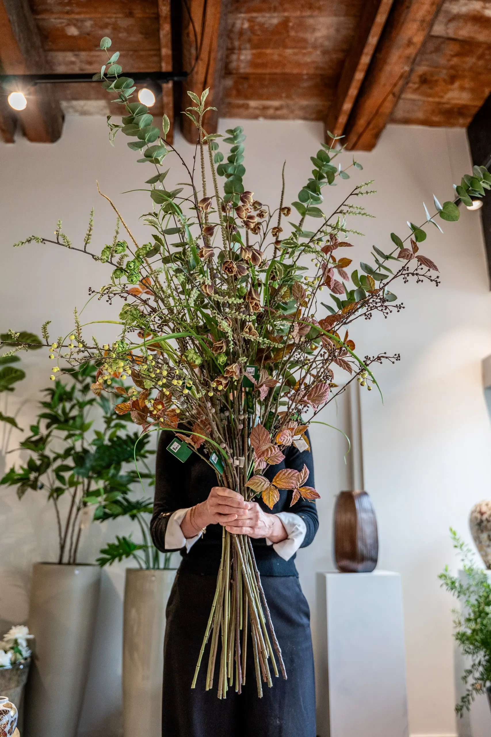 De eigenaresse houdt een hoog boeket van kunsttakken met eucalyptus en herfstblad voor zich, onder een houten balkenplafond