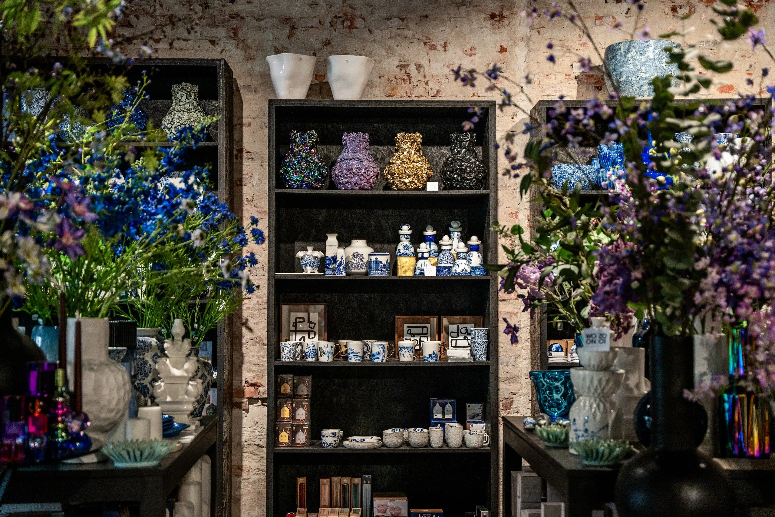 Een zwarte kast vol Delfts blauw servies en vaasjes, met blauwe en paarse bloemen ernaast, voor een oude bakstenen muur