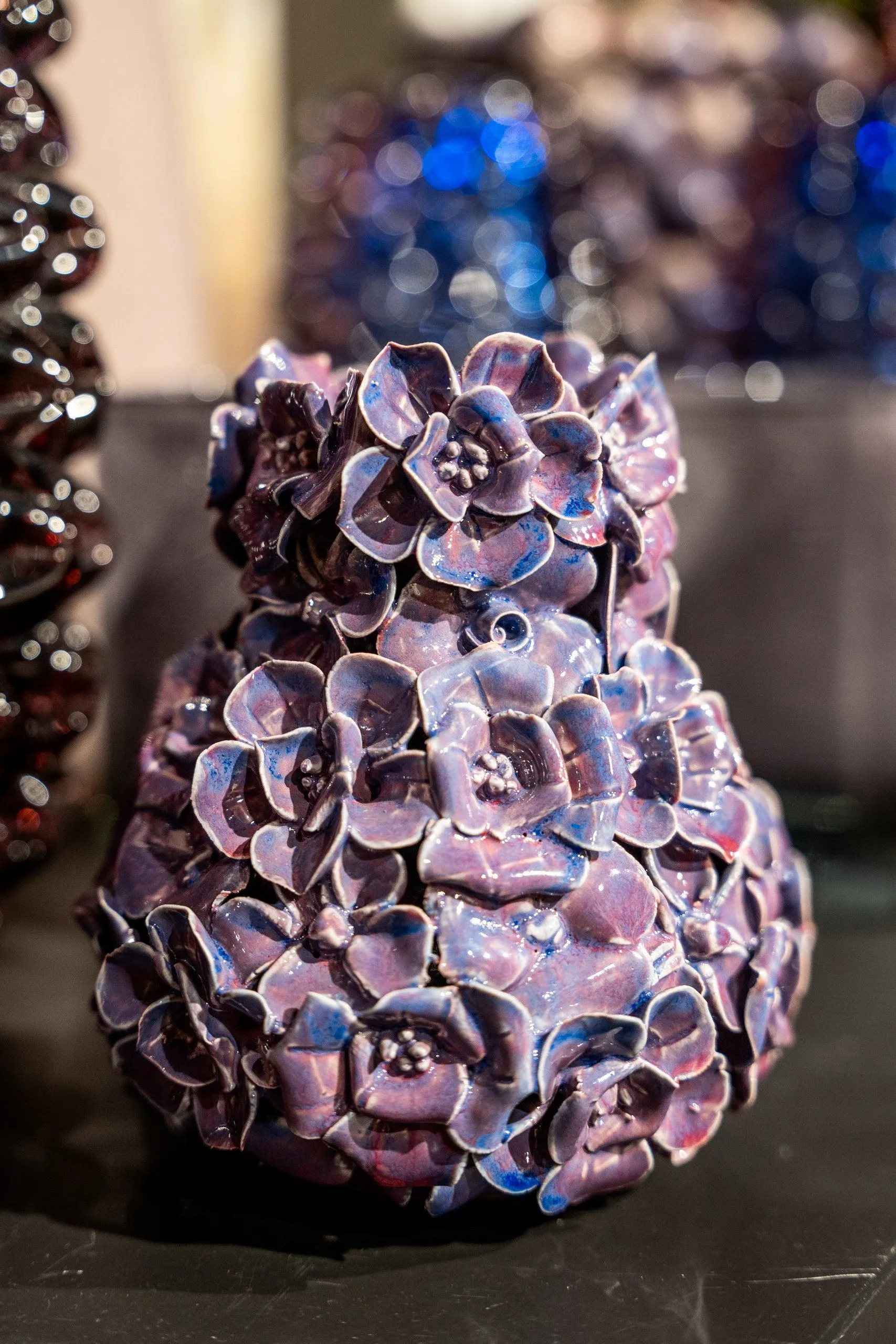 Een vaas vol keramieken bloemen in paars en roze, op een donkere tafel, met blauwe glazen vazen erachter