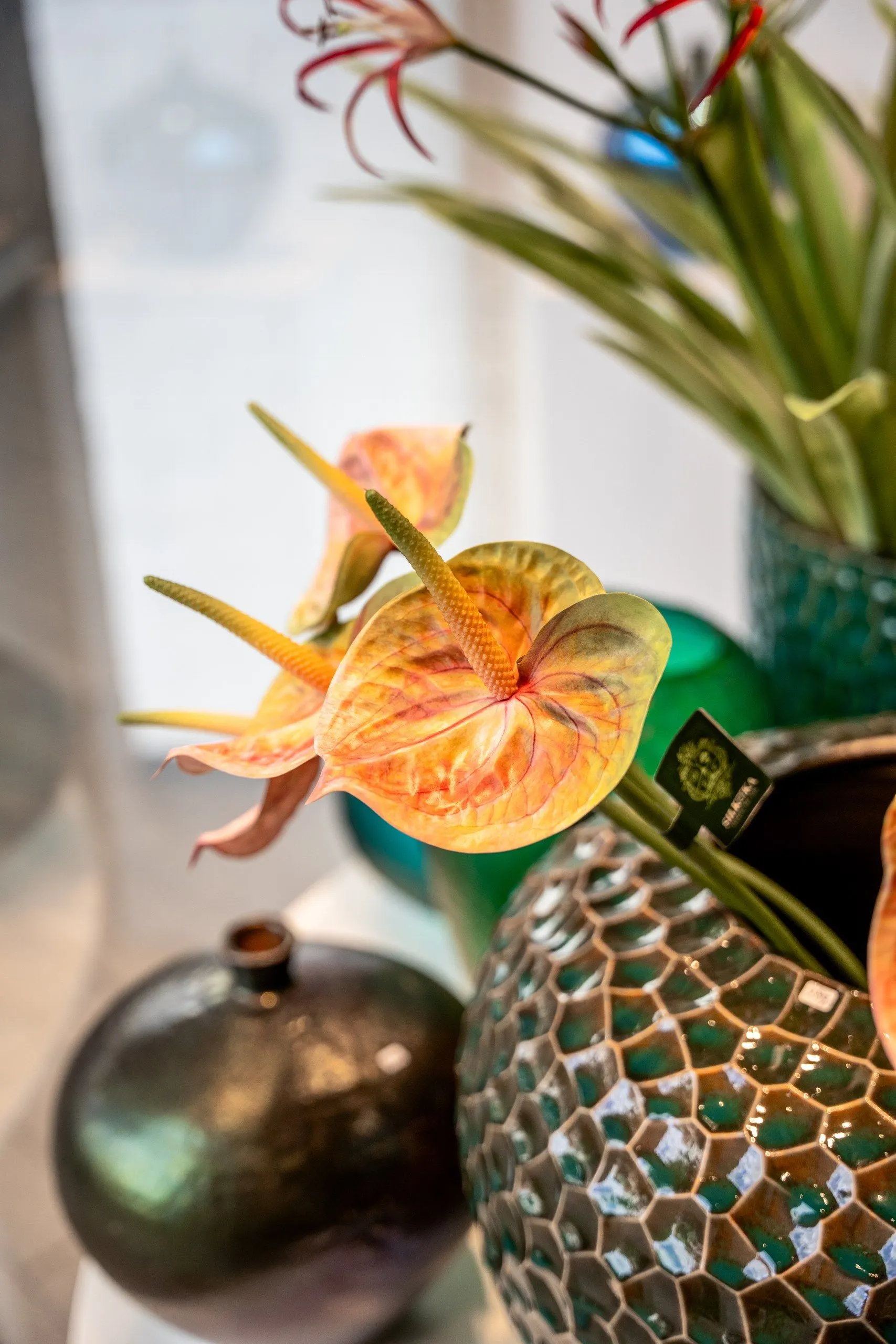 Een oranje anthurium boven een vaas met groen mozaïek en een bolle donkere vaas