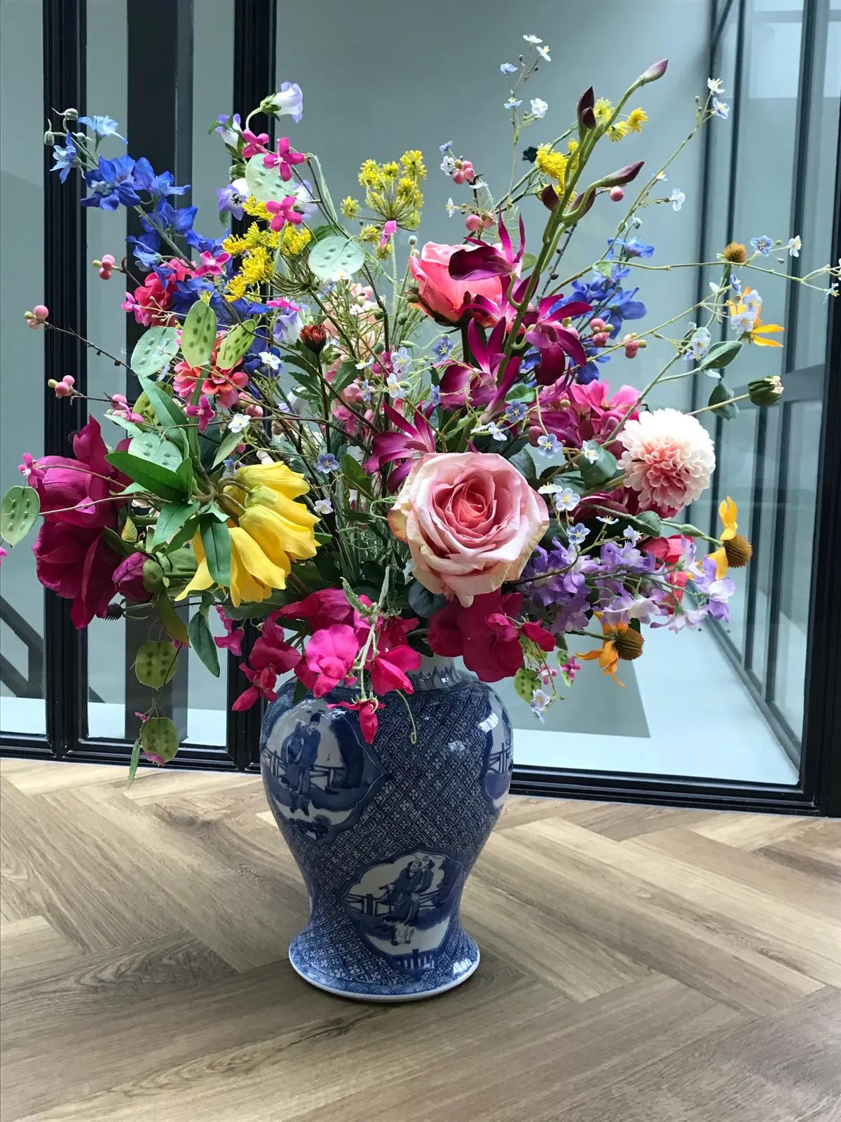 Kleurrijk boeket met een zalmroze roos, fuchsiaroze en gele bloemen, blauwe ridderspoor en judaspenning in een blauw-witte Delftse pot op een visgraatvloer