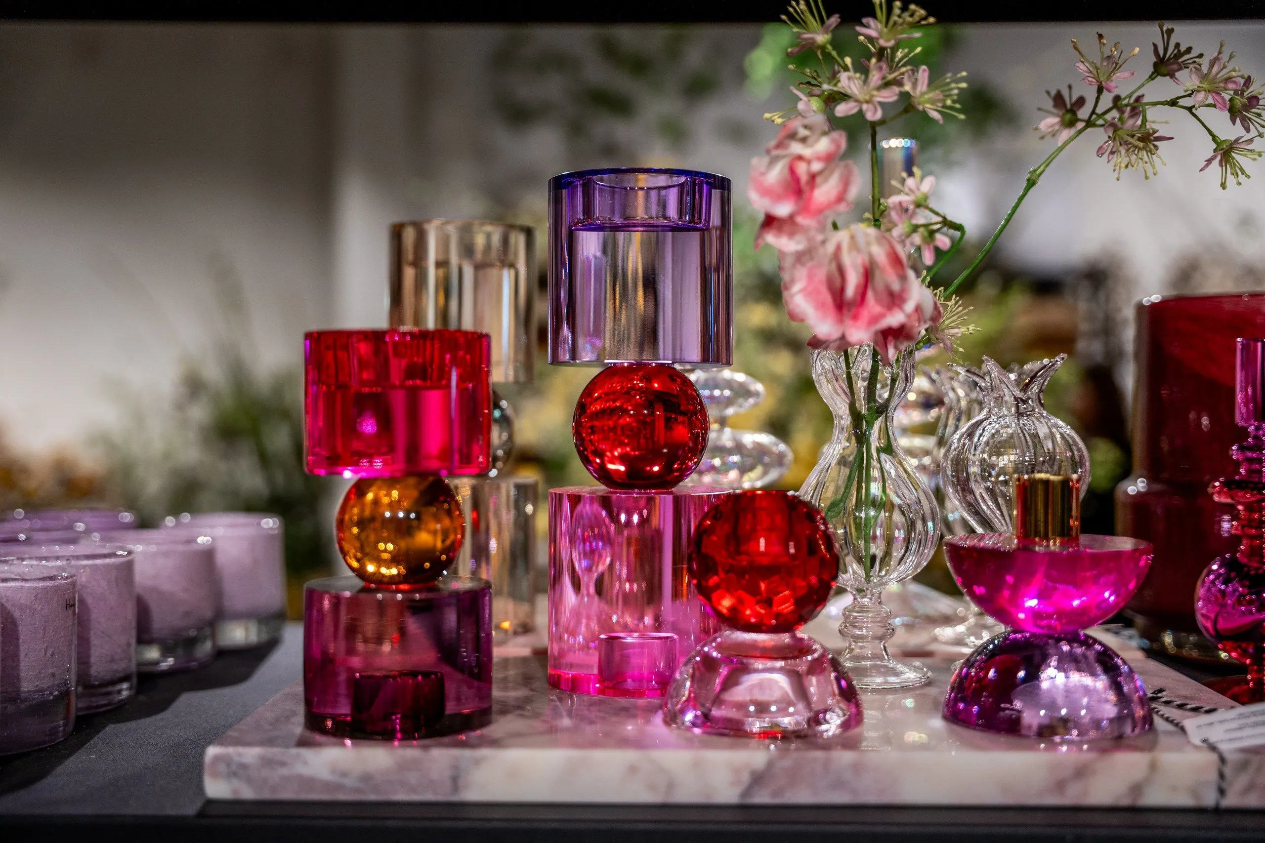 Kandelaars van gekleurd glas in roze, rood, paars en amber op een marmeren plateau, met een vaasje roze bloemen erachter