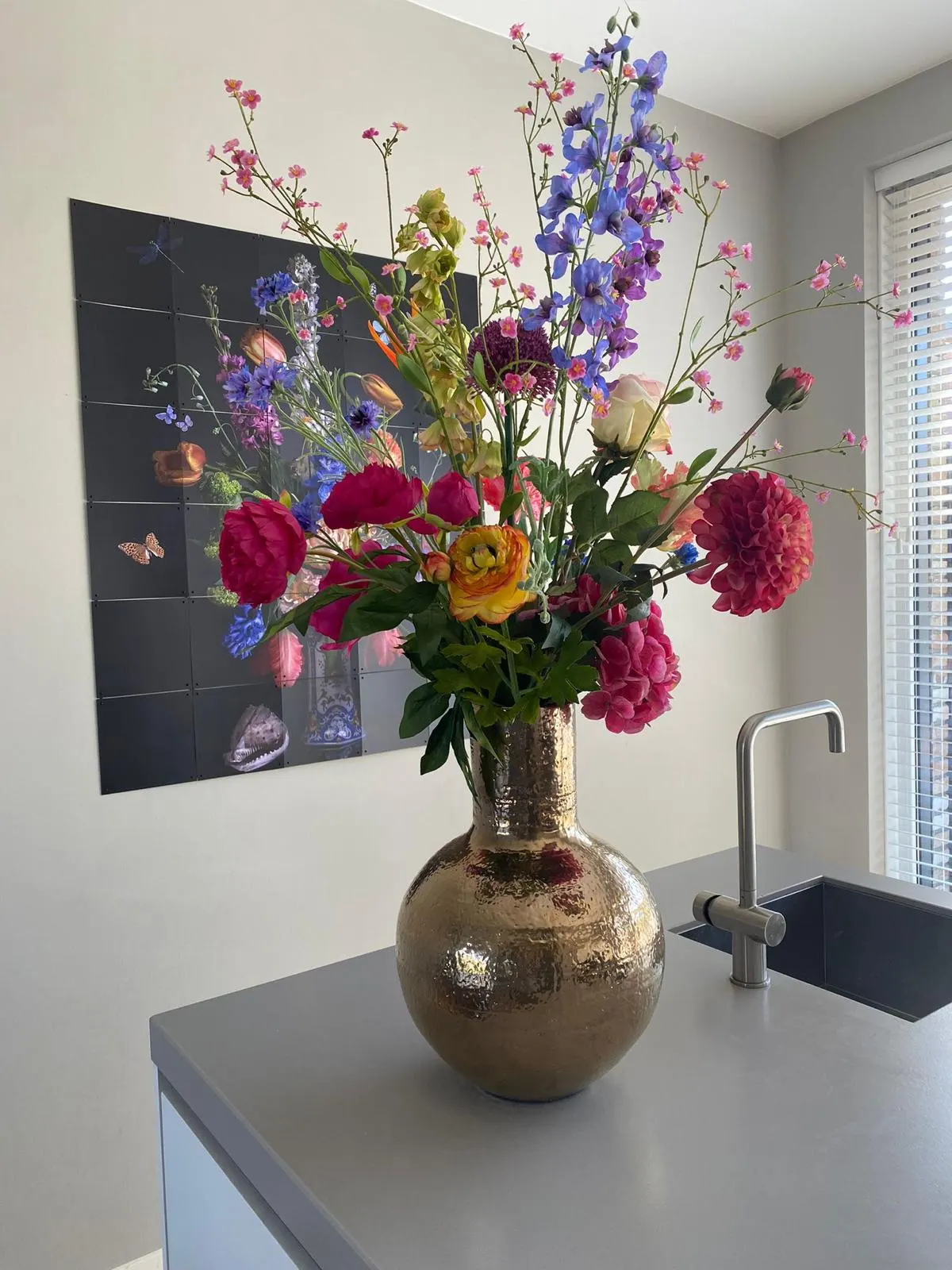Boeket met fuchsiaroze dahlia's en ranonkels, blauwe ridderspoor en roze takjes in een bolle gehamerde zilveren vaas op een grijs keukenblok