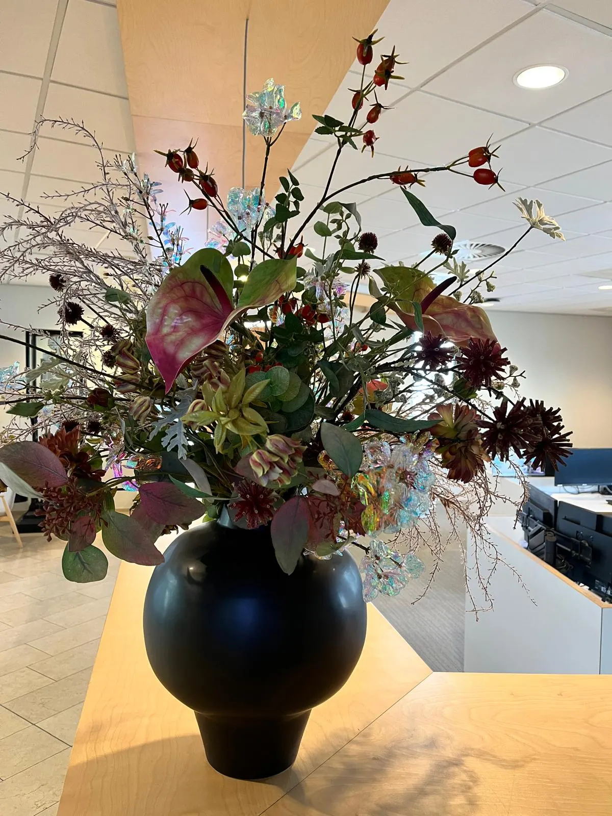 Herfstboeket met donkerrode dahlia's, roze-groene anthuriums, rozenbottels en parelmoerglanzende bloemen in een bolle zwarte vaas op een houten balie, in een kantoor