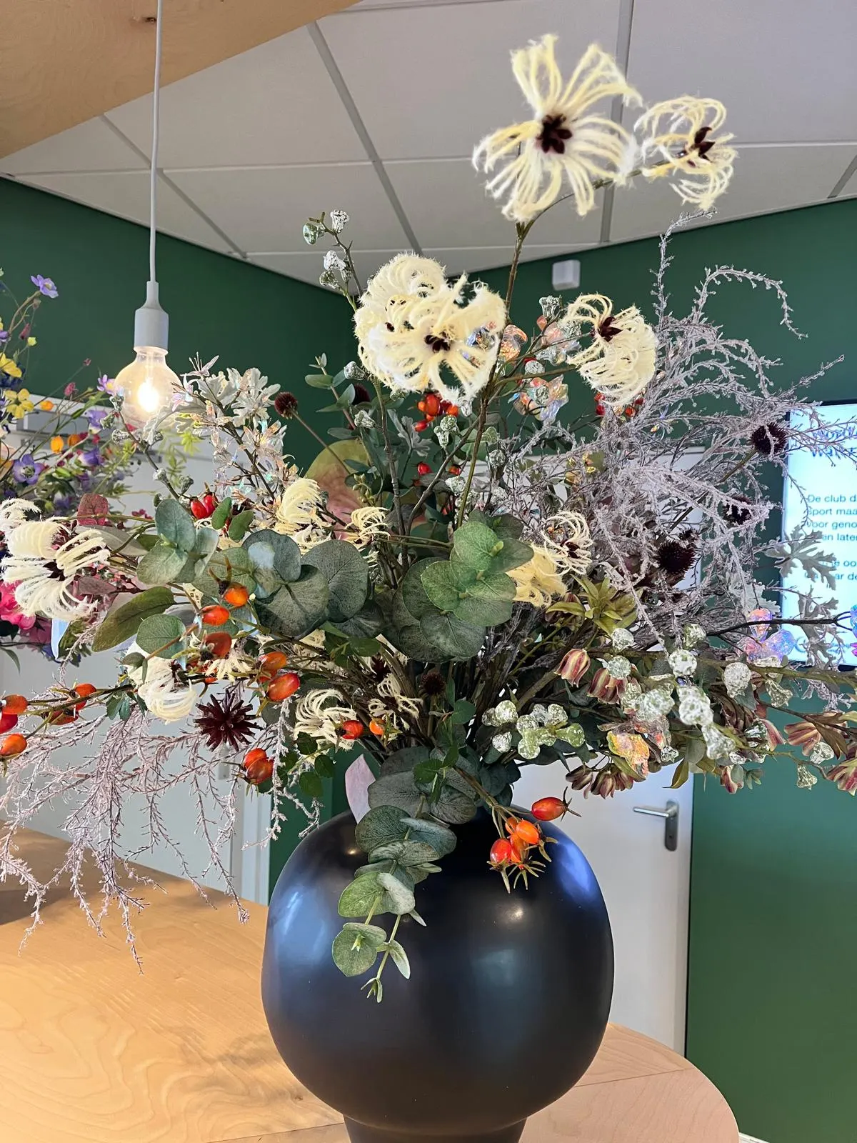 Boeket van crèmewitte bloemen met rafelige blaadjes, eucalyptus, rozenbottels en roze pluimen in een bolle zwarte vaas, in een kantoor met een groene wand