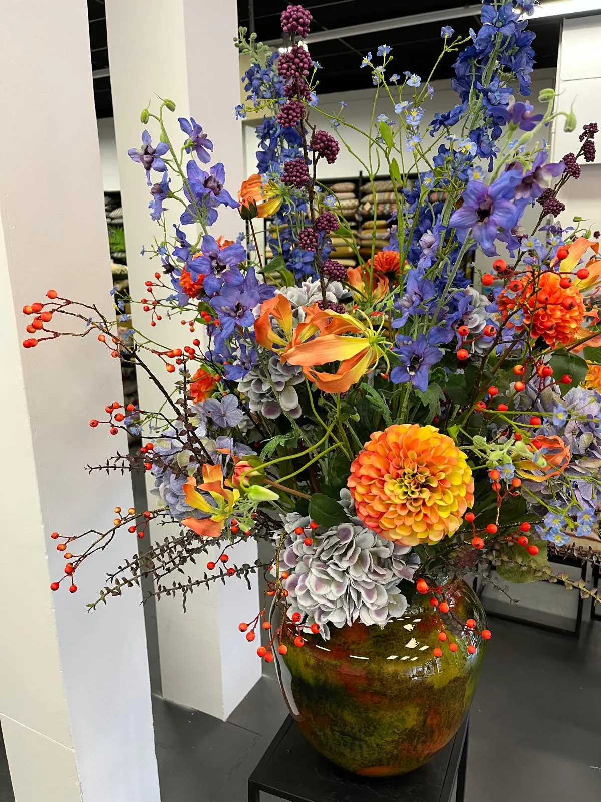 Groot boeket met blauwe ridderspoor, een oranje dahlia, gloriosa's, takken met oranje besjes en grijsblauwe hortensia in een groen-oranje geglazuurde vaas, tussen witte pilaren