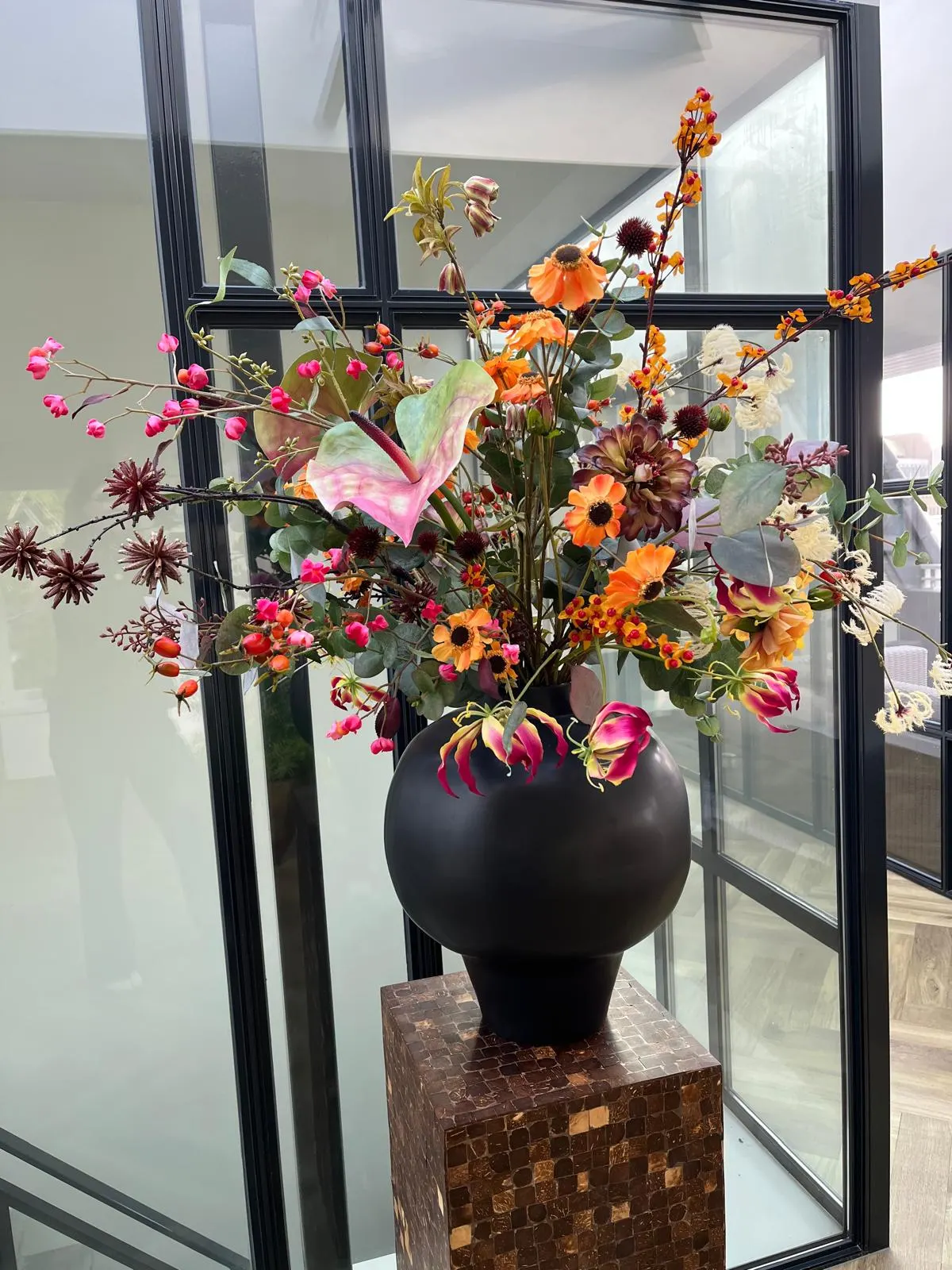 Boeket met oranje bloemen, gloriosa's, een roze anthurium en donkere distelbollen in een bolle zwarte vaas op een zuil van kokosmozaïek, voor een glazen wand met zwarte kozijnen