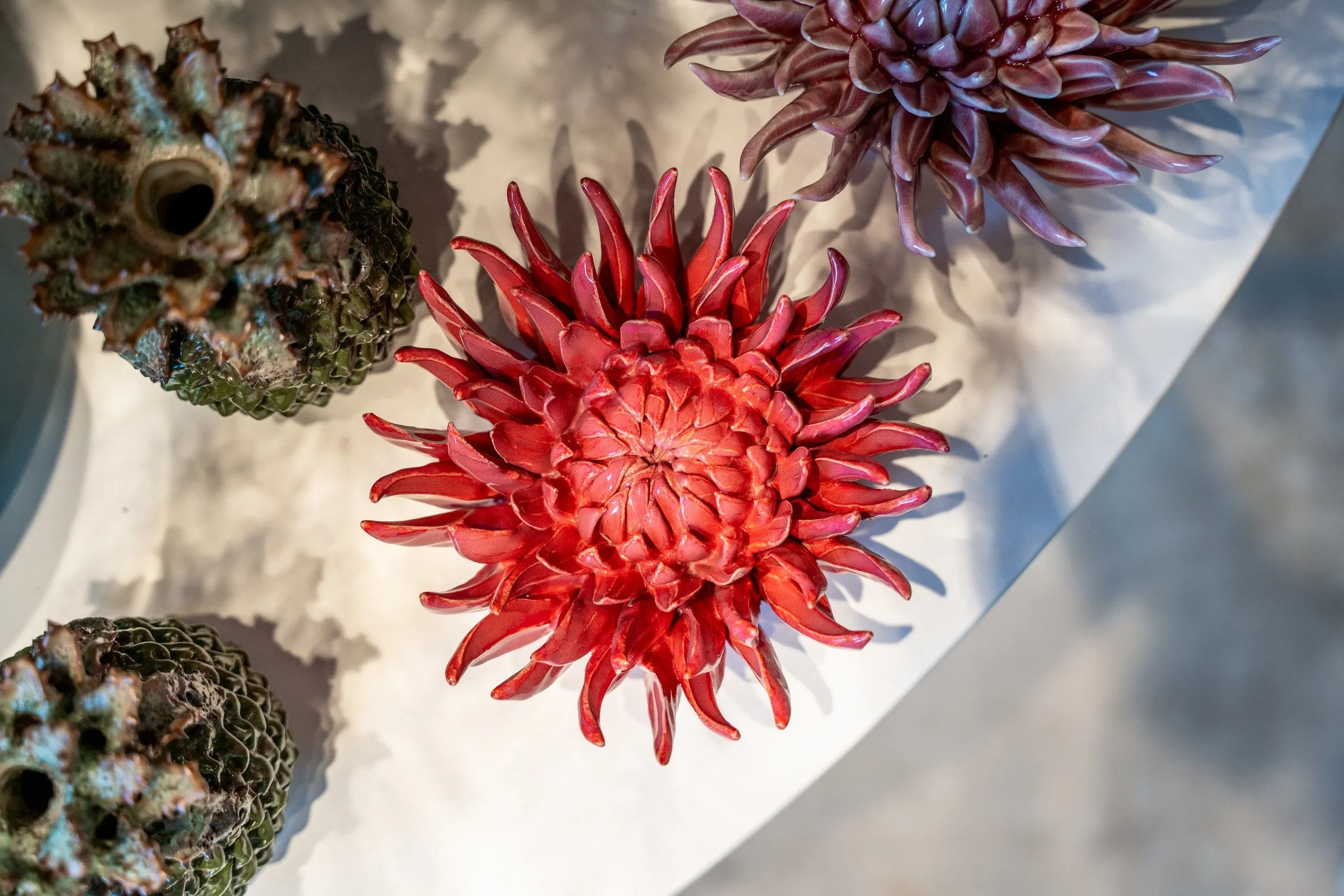 Bloemen van keramiek in rood en paars en twee bolle potjes, van bovenaf op een witte tafel in de zon