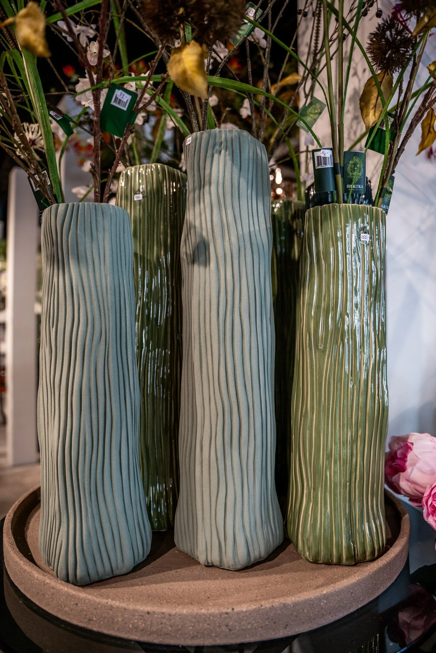 Drie hoge vazen met ribbels in grijsgroen en groen op een ronde houten schaal, met takken erin