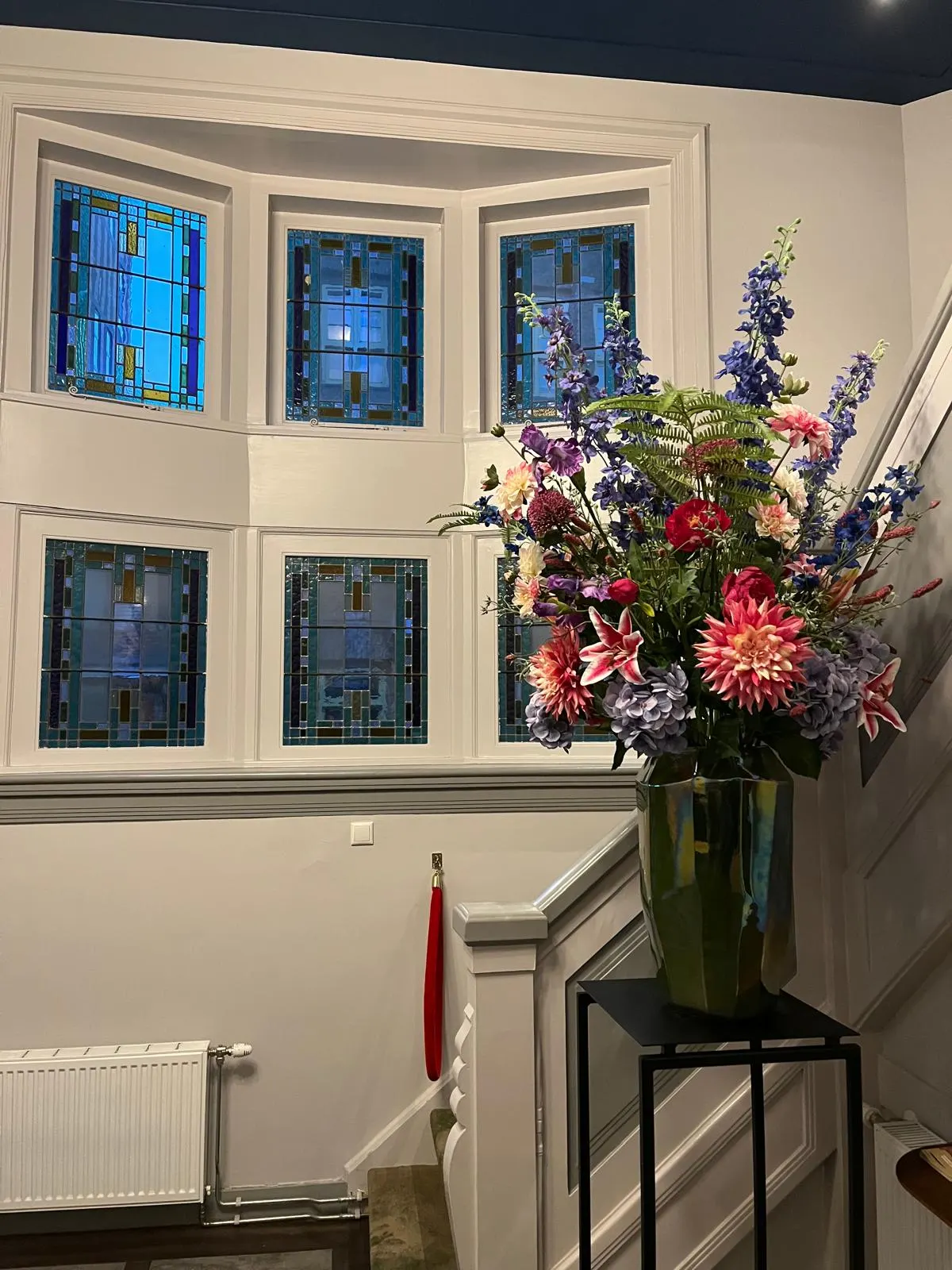 Boeket met blauwe ridderspoor, roze dahlia's en lelies in een glanzende donkere vaas op een zwart tafeltje, in een trappenhuis met zes glas-in-loodramen