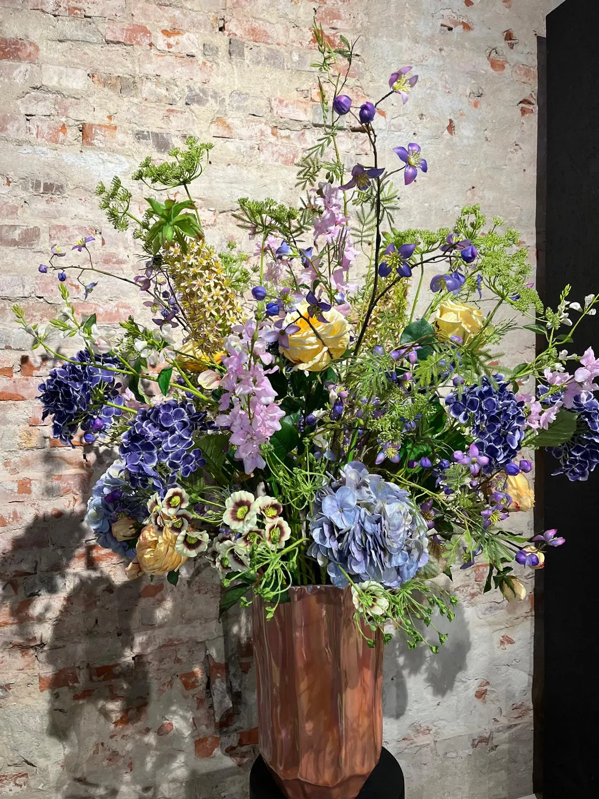 Weelderig boeket van blauwe en paarse hortensia's, lila ridderspoor, gele rozen en groene schermbloemen in een koperen vaas met facetten, voor een bakstenen muur