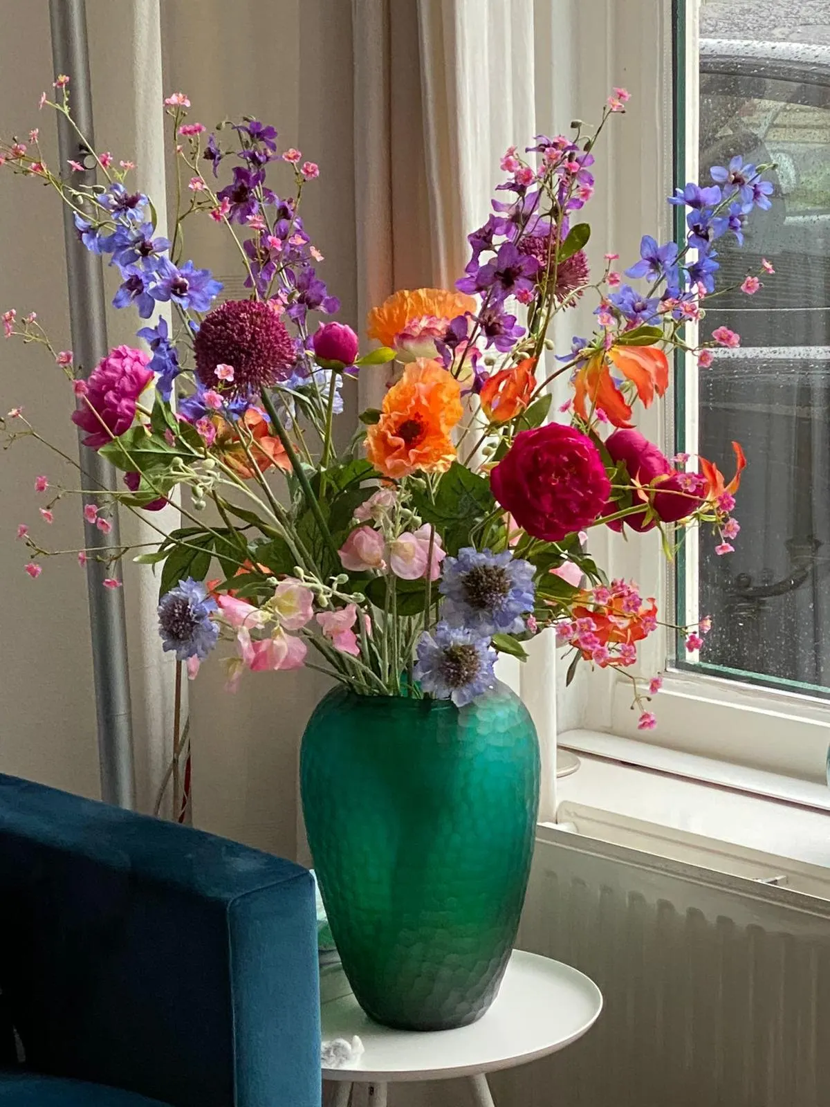 Boeket met oranje ranonkels, paarse ridderspoor, blauwe scabiosa's en roze lathyrus in een groene glazen vaas met facetten, op een wit bijzettafeltje bij het raam, naast een blauwe fluwelen stoel