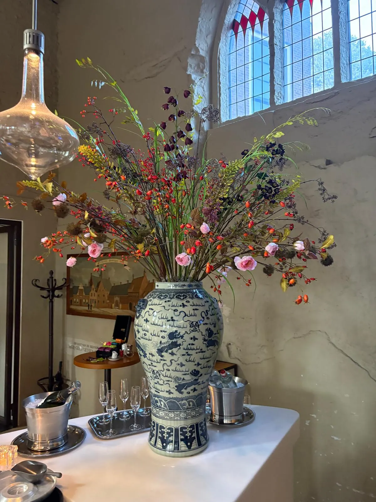 Hoge, losse schikking van takken met oranje rozenbottels, roze roosjes en donkerpaarse bloemen in een grote blauw-witte Chinese vaas, op een witte bar met een champagnekoeler, onder een glas-in-loodraam