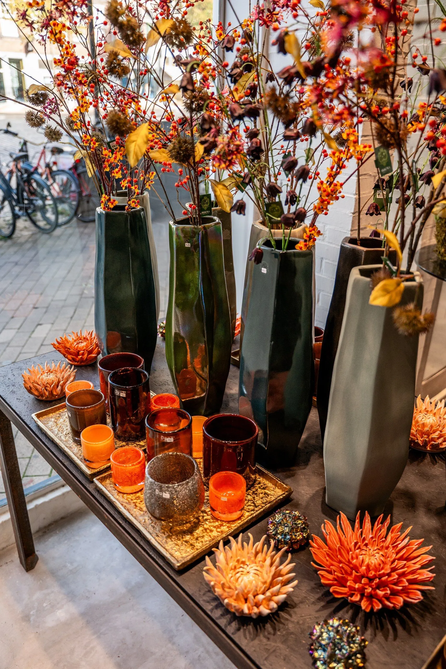 Herfsttakken met oranje besjes in hoge groene vazen voor het winkelraam, met oranje en amberkleurige kaarsglazen op gouden dienbladen en keramieken bloemen op de tafel
