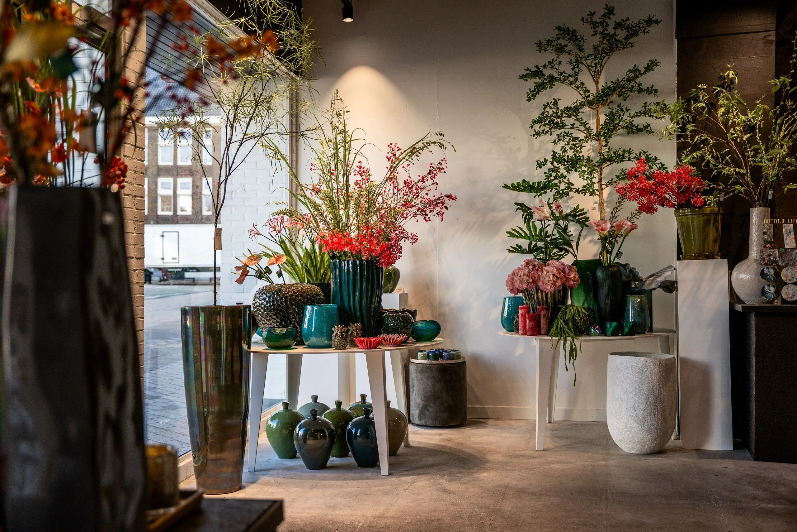 Hoek van de winkel bij het raam: groene en blauwe vazen op witte tafeltjes, rode bloemen en jonge boompjes, met de straat achter het glas